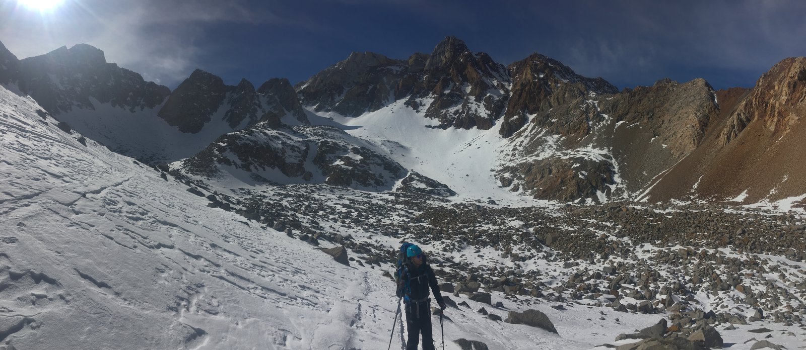 View of rocky but snow-covered mountain terrain. A skier is in the foreground. The sun is shining.