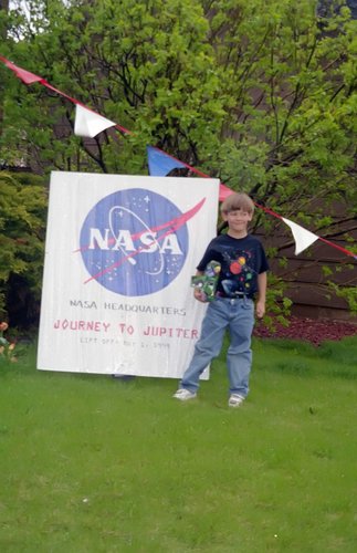 Young Tanner Harms standing in front of a sign that reads "NASA Headquarters, Journey to Jupiter. Lift off: May 1, 1999"