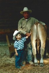 An old man wearing a cowboy hat holding the arm of a young child, also wearing a cowboy hat, who is touching a four-legged animal facing away from the camera.