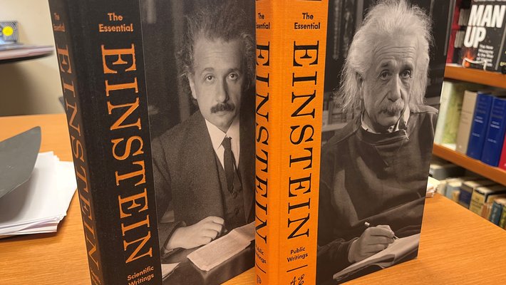 2 books, uprirght on a desk, bookshelf in background. Books are The Essential Einstein. Scientific Writings cover is of youthful Einstein. The Public Writings cover is a photo of an elderly  Einstein.