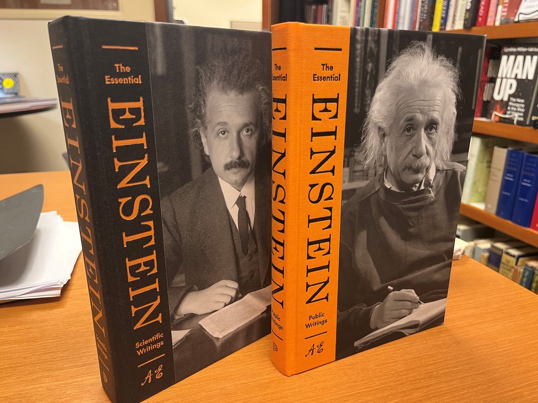 2 books, uprirght on a desk, bookshelf in background. Books are The Essential Einstein. Scientific Writings cover is of youthful Einstein. The Public Writings cover is a photo of an elderly Einstein.