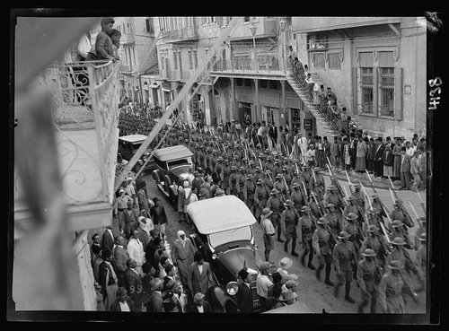 Black and white photo taken from a first floor balcony or window. Image is of British troops marching down street in Jerusalem with bayonets, alongside cars. Local people line the street observing the scene.