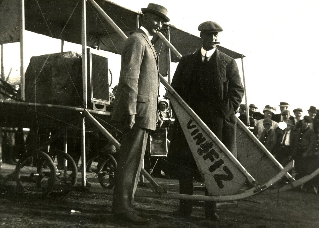 Pilot Cal Rodgers and an unidentified man stand in front of Rodgers's biplane, marked with an advertisement for "Vin Fiz," with a crowd of onlookers in the background.