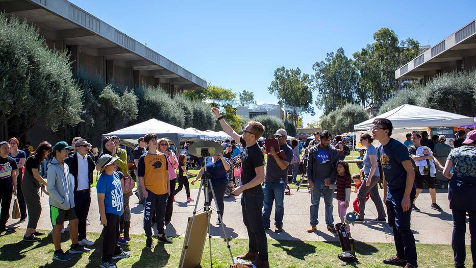 A crowd gathers to watch a person who is holding a stick with a flaming marshmallow at a 2019 Science for March event on Beckman Lawn on Caltech's campus.