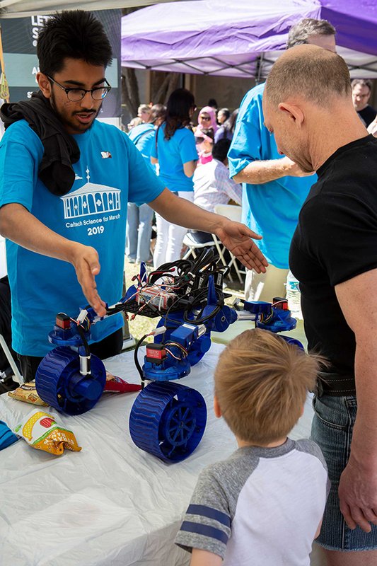 A person in a blue Caltech Science for March 2019 shirt gestures over a table containing a four-wheeled blue rover robot as a child and adult look at the robot. A crowd is visible under canopy tents behind the table.