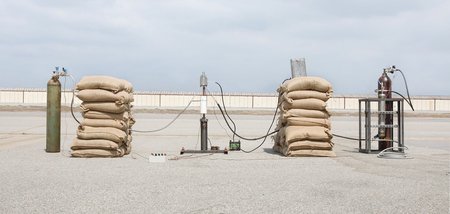 A sculptural installation consisting of a simple tripod rocket launcher with piles of sandbags on either side, and liquid fuel tanks on either side of the sandbags. The tanks and the launcher on the tripod are connected with many cables.