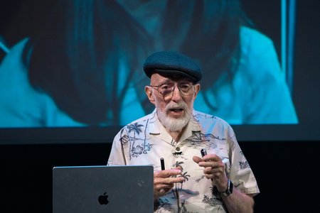 Kip Thorne, in a beret and button-down shirt, gestures as he speaks near a podium