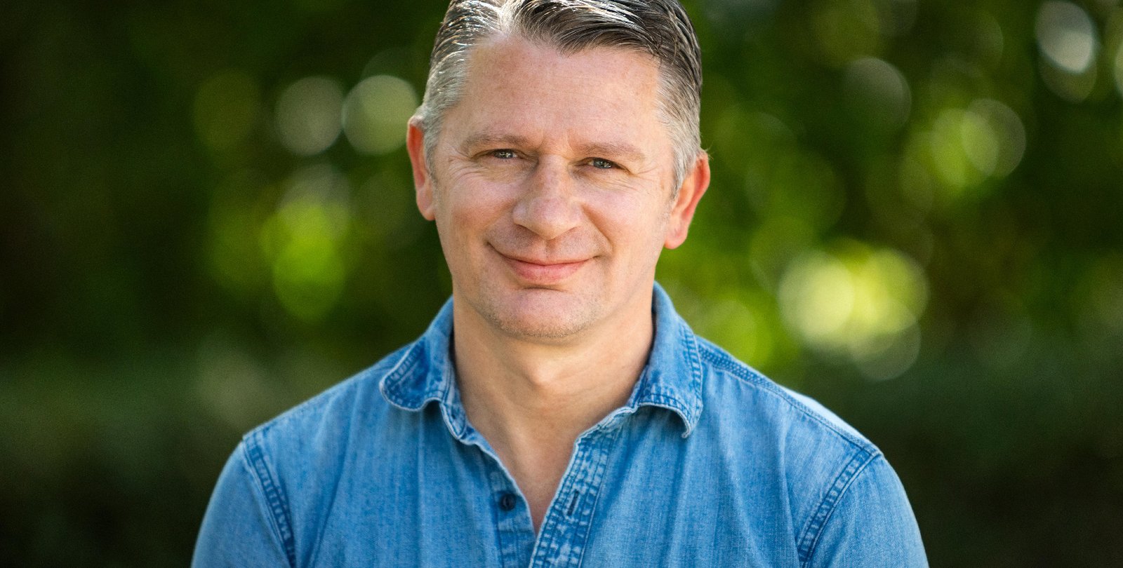 A portrait of Dean Mobbs. He wears a chambray shirt and smiles at the camera.