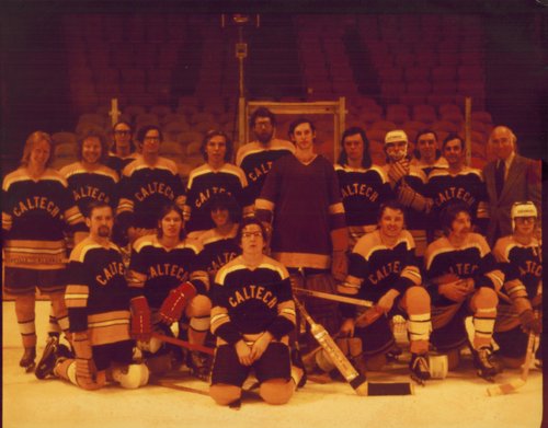 An old discolored photograph of a a hockey team wearing uniforms, some holding sticks, on an ice rink. Stadium-style seating can be seen behind them.