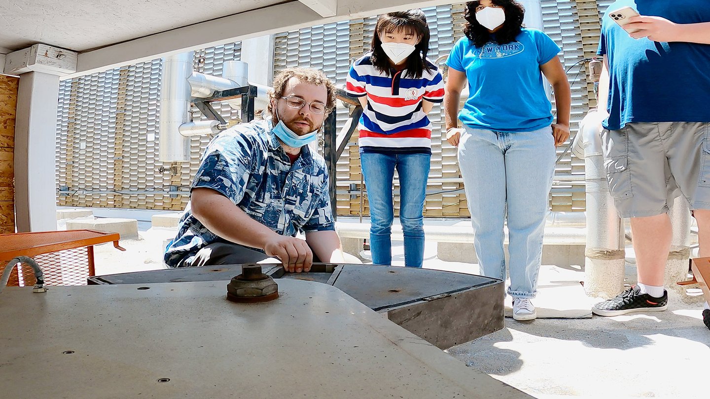 Four people are on a flat, white asphalt rooftop, three standing and one squatting in the foreground touching a large wedge-shaped metal object. Two of the three standing watch intently and the third looks at a phone. Pale blue sky is visible through a mid-century metal barrier.