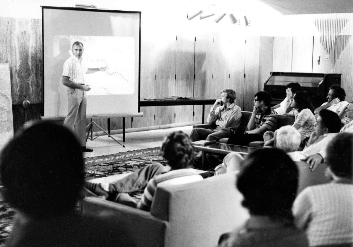 A black and white photo of a man lecturing before a projector screen to a class full of male students in 1970.