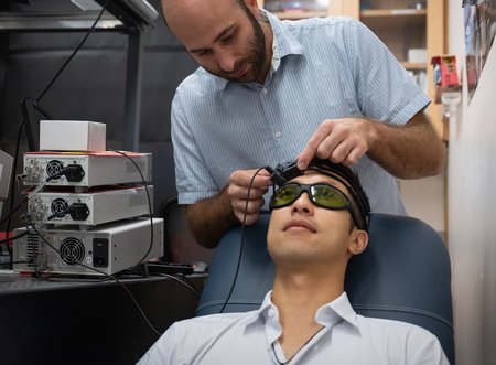 One man is putting a headset-based device on another man who is sitting in a chair in front of him.  The man in the chair is wearing sunglasses