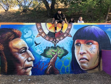 Mural painting on a wall with an indigenous man in profile on the left, a tree in the center, the sun above, and an indigenous girl in 3/4 profile on the right. A boy is sitting on top of the wall the mural is on, and two more children are behind the wall with their faces showing.