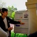 A young man wearing a white t-shirt and jacket points out something on a poster to two onlookers.