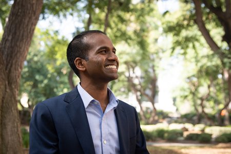 A portrait of Karthish Manthiram. He wears a button-down shirt and blazer and smiles. Trees are in the background.