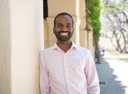 A man with brown skin and a beard wearing a pink button up shirt smiles warmly at the camera while leaning against a pillar.