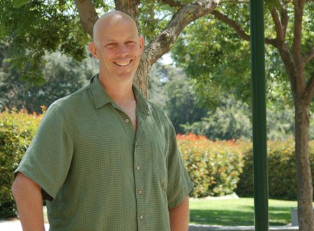 A man wears a green button down shirt and smiles for a photo outdoors