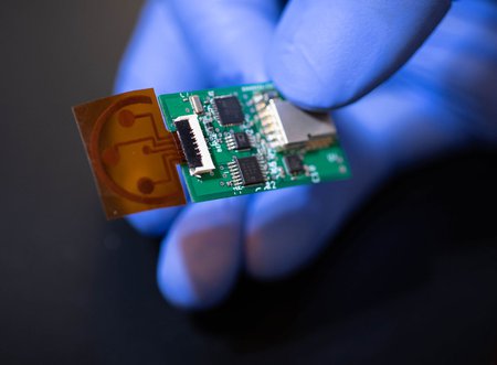A hand wearing a blue rubber glove holds a small sensor that consists of a circuit board and a thin piece of plastic film.