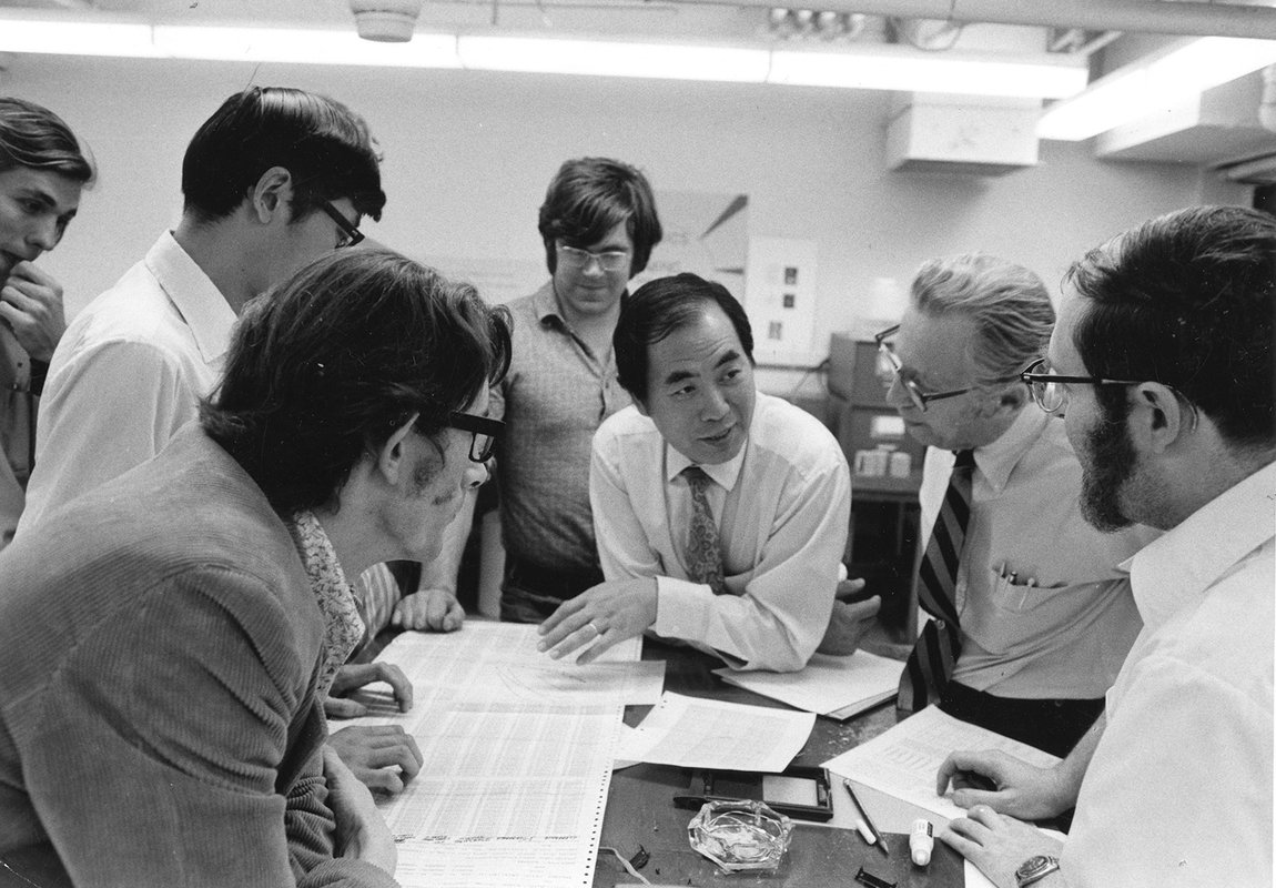 A man wearing a shirt and tie appears at the center of this black and white photo. He leans on a table surrounded by students who are listening to him as he talks.