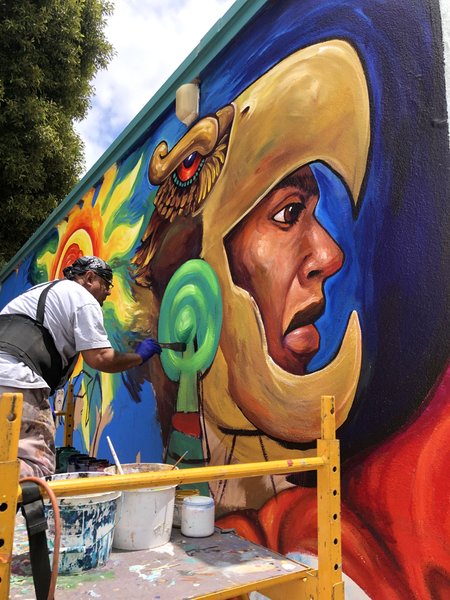 A man on scaffolding is painting a mural of a face wearing a bird mask in profile.