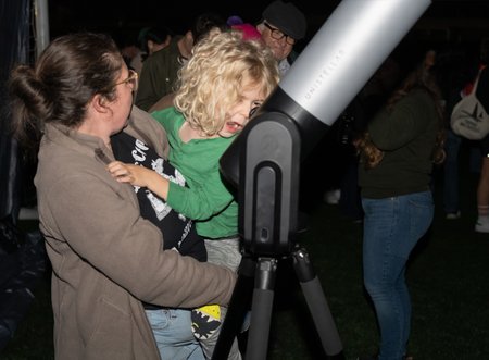 A woman holding a child with curly blonde hair who is leaning in to look into a telescope's eyepiece