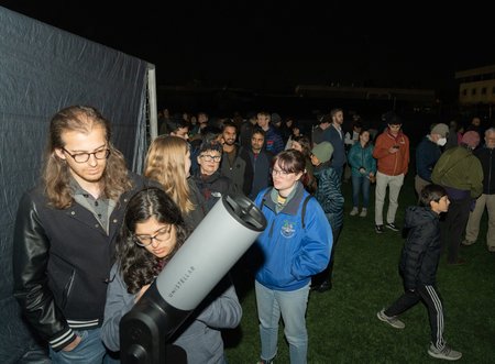 A crowd of people stands in line behind a telescope in the dark, illuminated by a camera flash