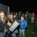 A crowd of people stands in line behind a telescope in the dark, illuminated by a camera flash