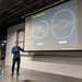 Cameron Hummels points to a projector screen with an image of the solar system displayed