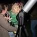 A woman holding a child with curly blonde hair who is leaning in to look into a telescope's eyepiece