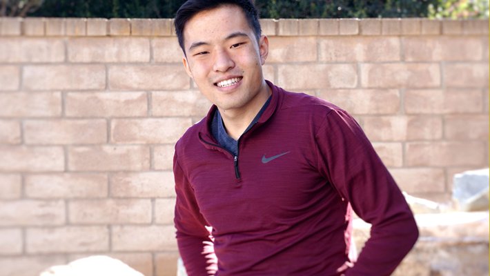 A portrait of chemistry student Andrew Zhou. He is sitting outside in front of a brick wall and some low hedges.
