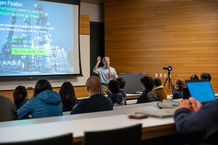 Jonas Peters speaks to an audience with a screen behind him displaying chemical equations