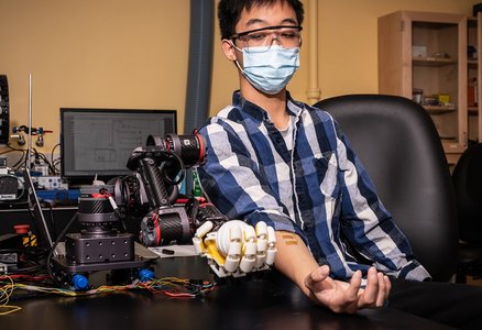 A human sits at a table flexing his hand. Sensors are attached to the skin of his forearm. A robotic hand next to him mimics his motion.