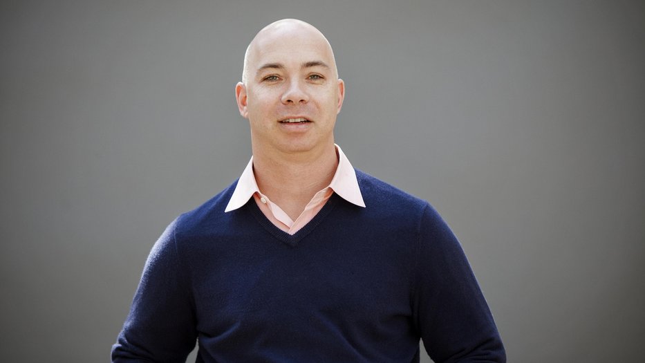 Portrait of person in in a dark blue sweater with a pink collared shirt on a gray background