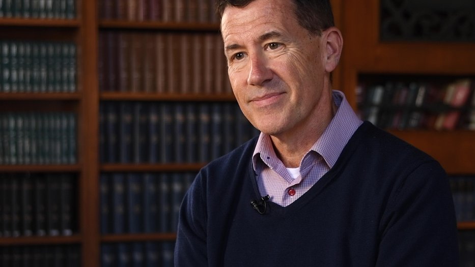 person in a sweater and collared shirt in front of a shelf full of books