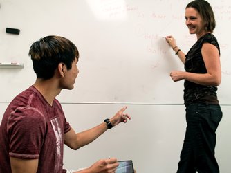 A woman writes on a white board and a seated student talks to her